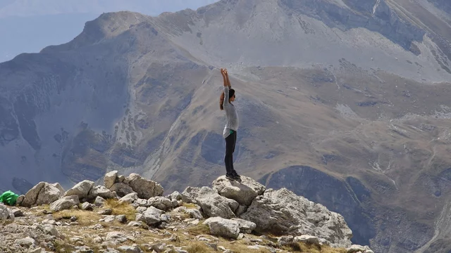 Ein Mensch steht auf einem Felsen und hebt beide Arme in die Luft. Im Hintergrund sind majestätische Berge und eine weite Landschaft zu sehen. Die Szenerie vermittelt ein Gefühl von Freiheit und Höhe.