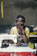 Eine lächelnde Frau mit Zopfhaar sitzt an einer Nähmaschine. Sie trägt ein buntes Kleid mit floralem Muster und Schmuck. Auf dem Tisch liegen Stoff und eine Tasche. Der Hintergrund ist unscharf, mit angedeuteten textilem Material.