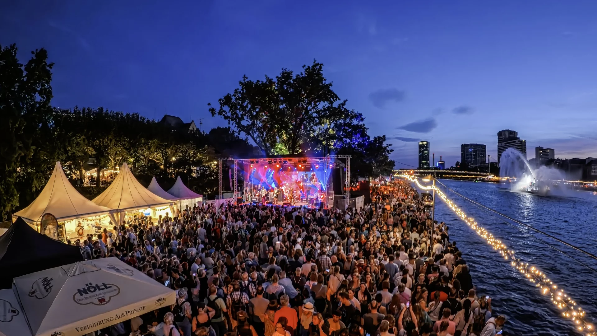 Ein belebtes Festival am Wasser bei Dämmerung. Eine große Menschenmenge genießt Live-Musik von einer belebten Bühne. Lichterketten und ein Wasserbrunnen schaffen eine festliche Atmosphäre. Hochhäuser und Bäume sind im Hintergrund sichtbar.