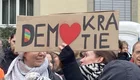 Eine Menschenmenge hält ein großes Plakat hoch, auf dem steht: "DEM♥KRA TIE". Das Herz ist rot und es gibt ein kleines Regenbogen-Symbol. Die Atmosphäre wirkt engagiert und demonstrativ, mit Personen im Vordergrund, die das Plakat betrachten.