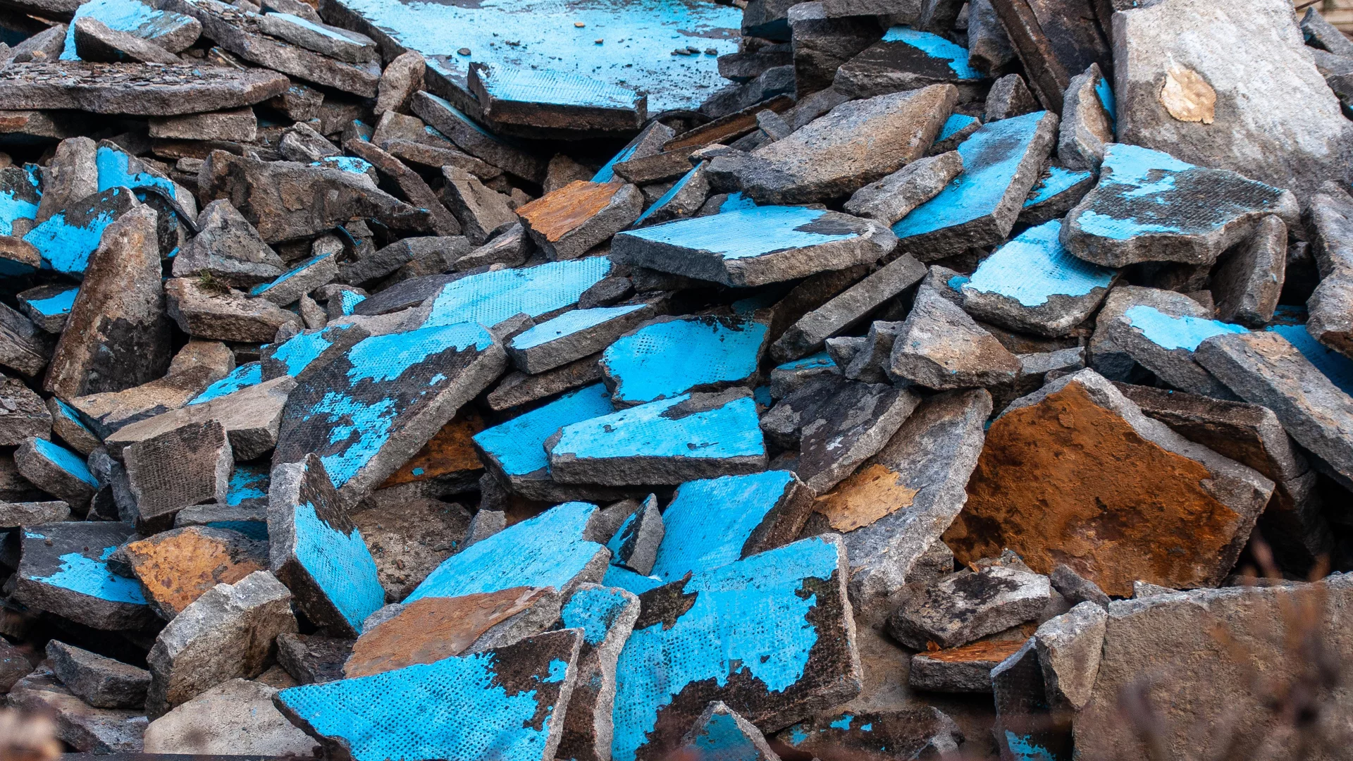Ein Haufen von abgebrochenen Betonplatten in verschiedenen Farben, vor allem in Blau und Grau. Die Platten sind unordentlich übereinander gestapelt und zeigen verschiedene Oberflächenstrukturen, die durch Abnutzung und Witterung geprägt sind.