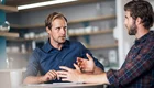 Zwei Männer sitzen an einem Tisch in einem hellen Raum und diskutieren. Der Mann links trägt ein blaues Hemd und wirkt nachdenklich, während der Mann rechts ein kariertes Hemd trägt und gestikuliert, als er spricht. Beide schauen sich aufmerksam an.