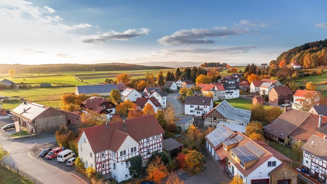 Eine malerische Landschaft mit einem kleinen Dorf im Herbst. Die Häuser sind von bunten, herbstlichen Bäumen umgeben. Im Hintergrund sind sanfte Hügel und ein klarer Himmel zu sehen. Die Szenerie vermittelt eine ruhige und idyllische Atmosphäre.