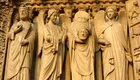 Reliefs an einer Kathedrale zeigen vier Figuren: links ein König, daneben ein Engel, der die Hände hebt. In der Mitte steht eine Figur mit einem Kopf in der Hand, rechts ein weiterer Engel. Die Figuren sind detailliert gestaltet und symbolisieren biblische Themen.
