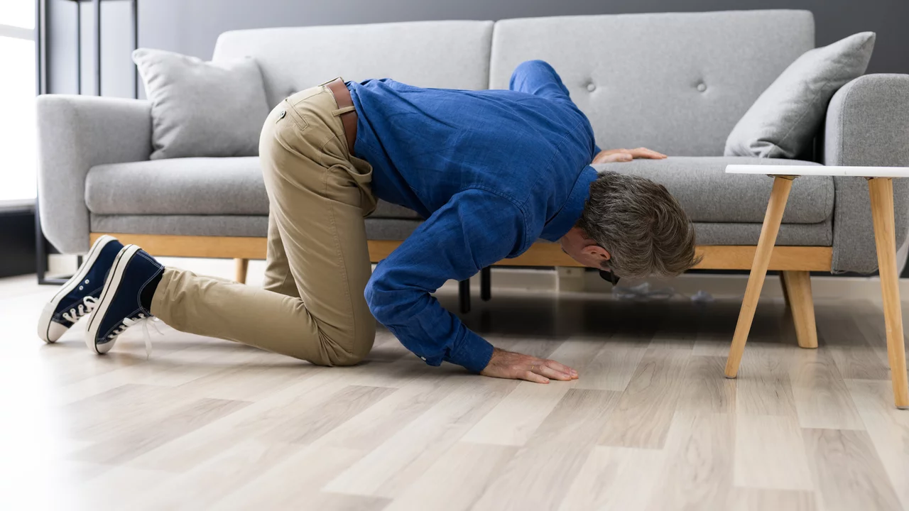 Ein Mann in einem blauen Hemd und khakifarbenen Hosen kniet auf dem Boden und schaut unter einem grauen Sofa nach. Er scheint nach verlorenem Gegenstand zu suchen, während das Wohnzimmer in einem modernen Stil eingerichtet ist.