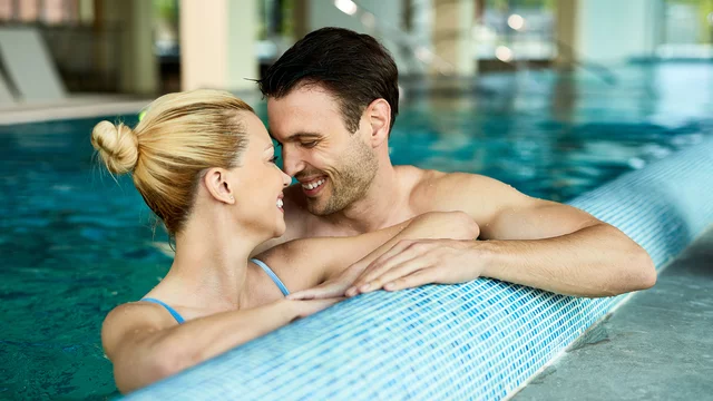 Ein glückliches Paar steht in einem Innenpool. Sie lächeln sich an und sind nah beieinander, wobei sie sich an der Poolkante stützen. Der Fokus liegt auf ihrer Verbindung und Freude miteinander.