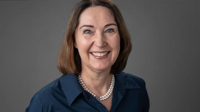 Eine lächelnde Frau mit schulterlangen, braunen Haaren trägt eine Perlencollier und ein dunkelblaues Hemd. Der Hintergrund ist neutral und unterstützt den Fokus auf ihr freundliches Gesicht.