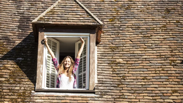 Eine fröhliche Person steht lächelnd an einem offenen Dachfenster und streckt die Arme der Sonne entgegen. Der Hintergrund zeigt ein Dach mit Ziegeln. Die Szene vermittelt ein Gefühl von Freude und Freiheit.