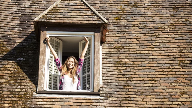 Eine fröhliche Person steht lächelnd an einem offenen Dachfenster und streckt die Arme der Sonne entgegen. Der Hintergrund zeigt ein Dach mit Ziegeln. Die Szene vermittelt ein Gefühl von Freude und Freiheit.