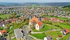 Eine luftige Aufnahme zeigt eine kleine Kirche mit rotem Dach, umgeben von Wiesen und Bäumen. Im Hintergrund erstreckt sich eine Stadt mit zahlreichen Wohnhäusern. Der Blick vermittelt ein Gefühl der Weite und grünen Landschaft.