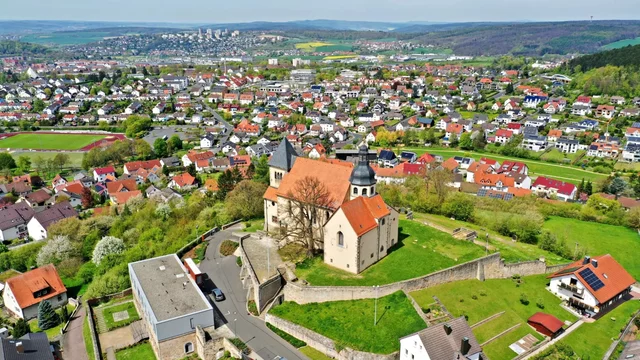 Eine luftige Aufnahme zeigt eine kleine Kirche mit rotem Dach, umgeben von Wiesen und Bäumen. Im Hintergrund erstreckt sich eine Stadt mit zahlreichen Wohnhäusern. Der Blick vermittelt ein Gefühl der Weite und grünen Landschaft.