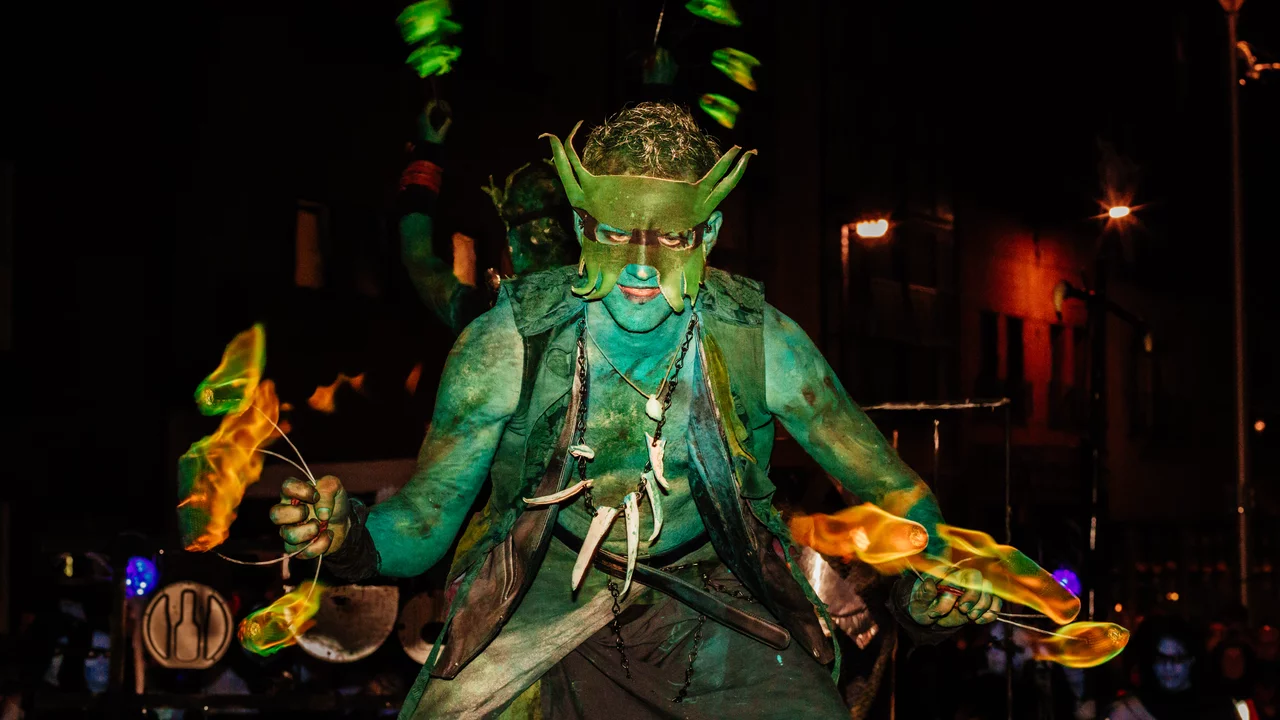 Ein künstlerisch geschminkter Performer mit grüner Körperfarbe steht im Vordergrund, während er mit brennenden Fackeln jongliert. Die Szene spielt nachts, beleuchtet durch städtische Lichter im Hintergrund, die eine festliche Atmosphäre schaffen.