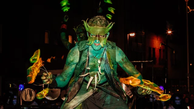 Ein künstlerisch geschminkter Performer mit grüner Körperfarbe steht im Vordergrund, während er mit brennenden Fackeln jongliert. Die Szene spielt nachts, beleuchtet durch städtische Lichter im Hintergrund, die eine festliche Atmosphäre schaffen.