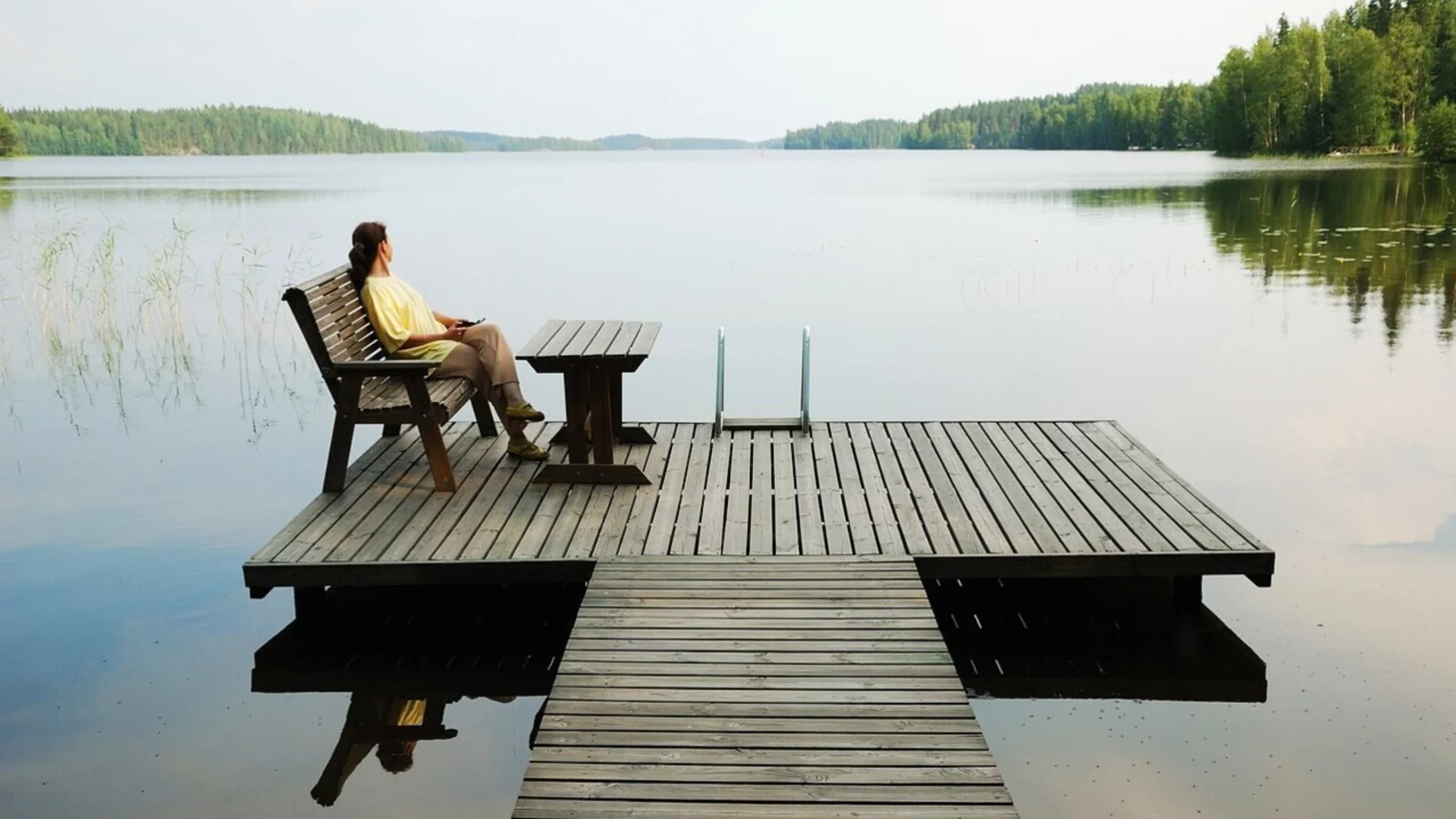 Eine Person sitzt auf einer Holzbank auf einem Steg, der über einen ruhigen See ragt. Die Umgebung ist von Wäldern umgeben, und die Stimmung ist friedlich mit einem klaren Himmel im Hintergrund.