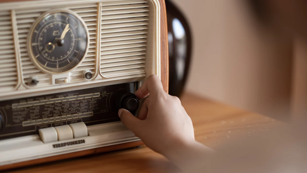 Eine Hand dreht an einem Regler eines retro Radioapparats. Der Radio hat ein Ziffernblatt und befindet sich auf einem Holztisch. Im Hintergrund sind unscharfe Möbel zu sehen.