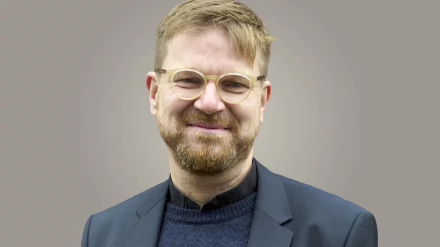 Ein Mann mit Brille und einem Bart lächelt in die Kamera. Er trägt einen dunkelblauen Pullover und einen grauen Blazer. Der Hintergrund ist einfarbig und neutral.