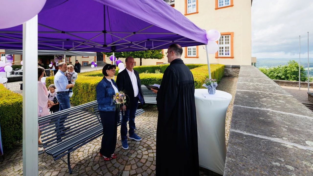 Eine Braut und ein Bräutigam stehen unter einem lila Zelt und halten einen Blumenstrauß. Ein männlicher Offiziant spricht mit ihnen. Im Hintergrund sind einige Gäste und eine historische Gebäudeumgebung zu sehen. Es ist ein schöner, heller Tag.