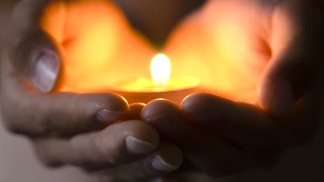 Zwei Hände halten eine kleine, brennende Kerze, deren warmes Licht sanft strahlt. Die Hände scheinen Schutz und Geborgenheit zu bieten, während die Kerze im Mittelpunkt steht und eine Atmosphäre der Ruhe und Besinnlichkeit vermittelt.