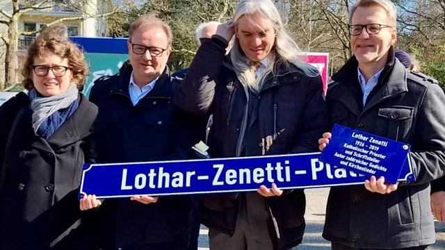 Vier Personen stehen zusammen und halten ein neues Straßenschild mit der Aufschrift „Lothar-Zenetti-Platz“. Die Szene findet im Freien statt, mit Bäumen im Hintergrund. Es scheint eine feierliche Zeremonie zur Enthüllung des Schildes zu sein.