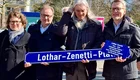 Vier Personen stehen zusammen und halten ein neues Straßenschild mit der Aufschrift „Lothar-Zenetti-Platz“. Die Szene findet im Freien statt, mit Bäumen im Hintergrund. Es scheint eine feierliche Zeremonie zur Enthüllung des Schildes zu sein.