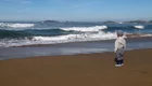 Ein kleines Kind steht am Strand und schaut auf die Wellen des Ozeans. Der Himmel ist klar und blau, und in der Ferne sind Blauen Hügel zu sehen. Der Sand ist golden und feucht von den umspülenden Wellen.