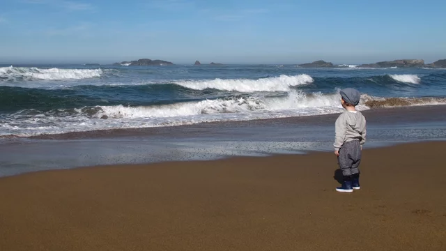 Ein kleines Kind steht am Strand und schaut auf die Wellen des Ozeans. Der Himmel ist klar und blau, und in der Ferne sind Blauen Hügel zu sehen. Der Sand ist golden und feucht von den umspülenden Wellen.