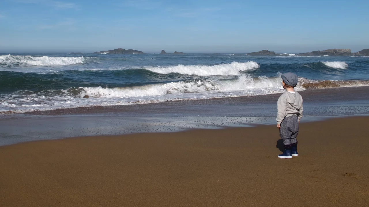Ein kleines Kind steht am Strand und schaut auf die Wellen des Ozeans. Der Himmel ist klar und blau, und in der Ferne sind Blauen Hügel zu sehen. Der Sand ist golden und feucht von den umspülenden Wellen.