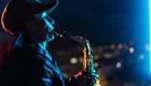 Eine Frau steht auf einem Dach und spielt Saxophon. Ihr Profil ist beleuchtet, und sie trägt eine Mütze. Im Hintergrund sind unscharfe Lichter einer Stadt bei Nacht zu sehen.