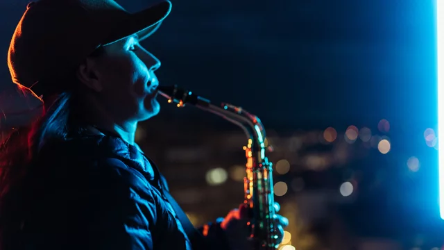 Eine Frau steht auf einem Dach und spielt Saxophon. Ihr Profil ist beleuchtet, und sie trägt eine Mütze. Im Hintergrund sind unscharfe Lichter einer Stadt bei Nacht zu sehen.