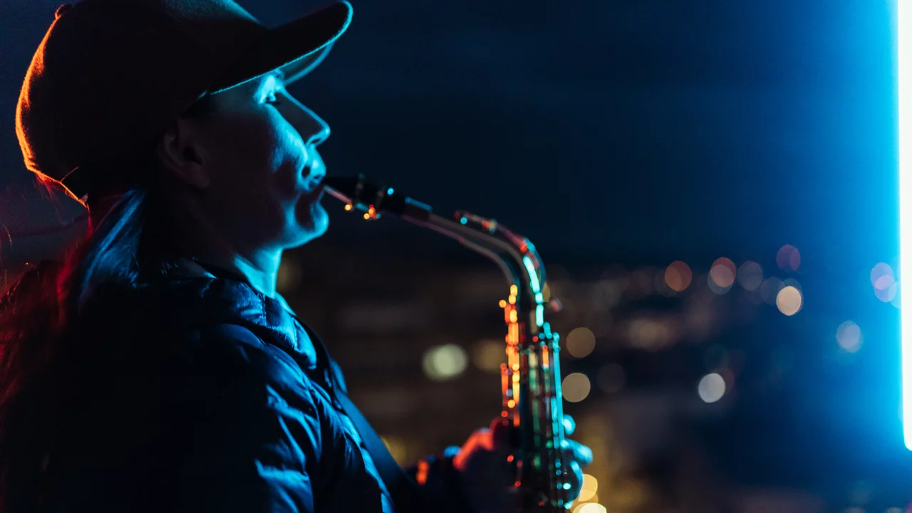 Eine Frau steht auf einem Dach und spielt Saxophon. Ihr Profil ist beleuchtet, und sie trägt eine Mütze. Im Hintergrund sind unscharfe Lichter einer Stadt bei Nacht zu sehen.