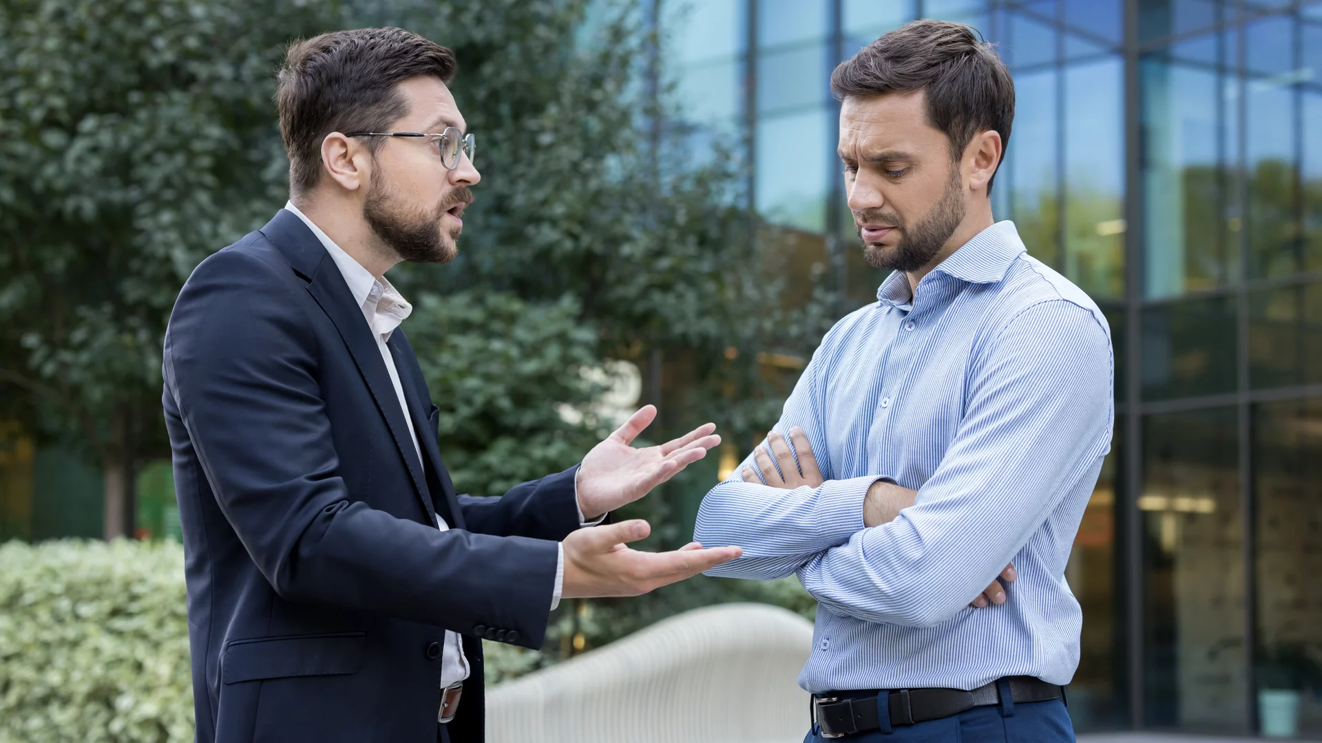 Zwei Männer stehen im Freien und führen ein angeregtes Gespräch. Der Mann links, im Anzug, gestikuliert und wirkt bemüht, während der Mann rechts, in einem Hemd, mit verschränkten Armen nachdenklich und skeptisch aussieht. Im Hintergrund sind Büsche und moderne Architektur sichtbar.
