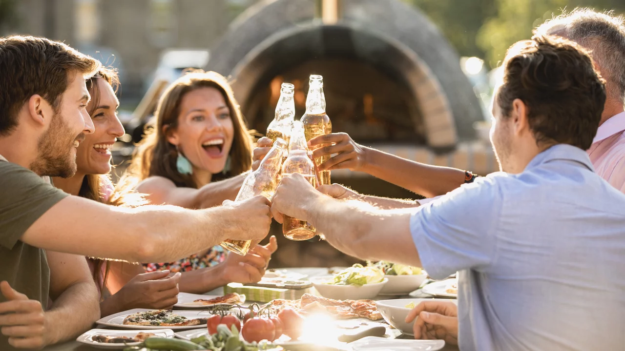 Fröhliche Menschen sitzen im Freien am Tisch und stoßen mit Getränken an. 
