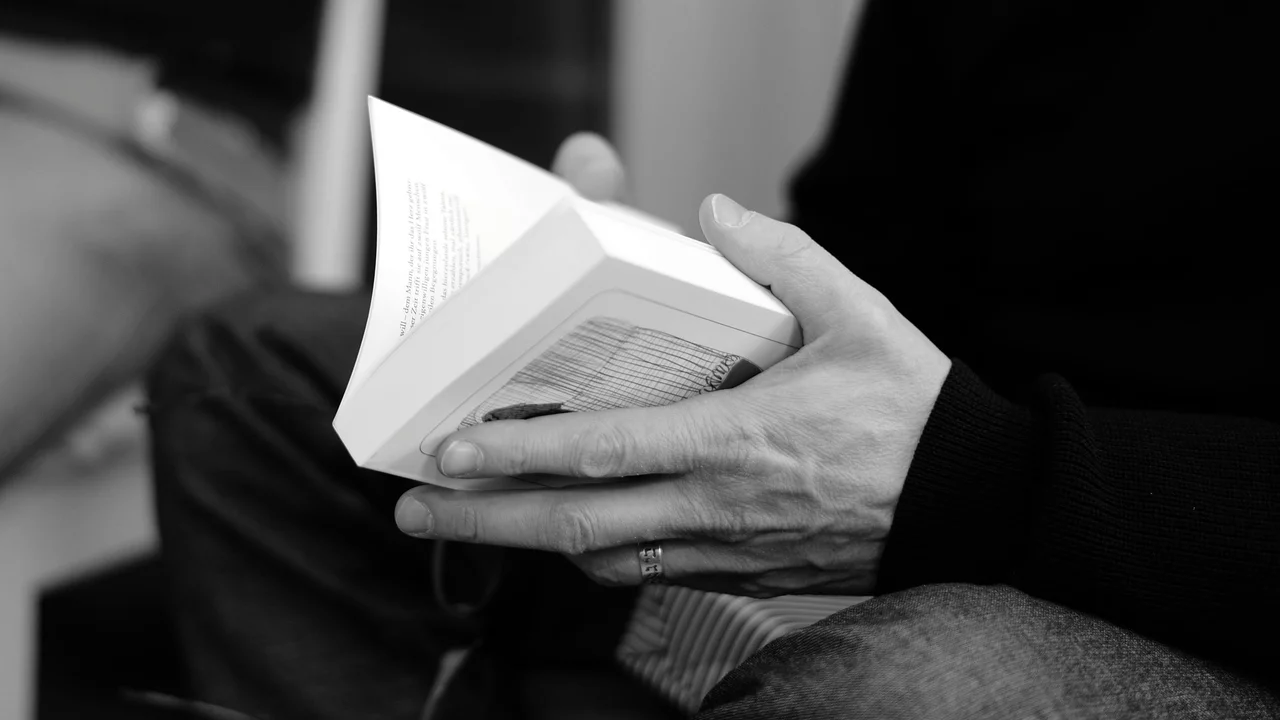 Eine Hand hält ein offenes Buch. Der Finger zeigt auf den Text. Das Bild vermittelt eine ruhige Lesesituation.