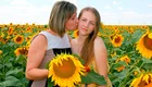 Zwei Frauen stehen in einem Feld voller Sonnenblumen. Eine Frau küsst die andere auf die Wange. Beide halten große Sonnenblumen in den Händen und lächeln. Der Himmel ist blau mit einigen Wolken.