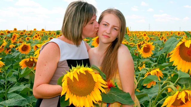 Zwei Frauen stehen in einem Feld voller Sonnenblumen. Eine Frau küsst die andere auf die Wange. Beide halten große Sonnenblumen in den Händen und lächeln. Der Himmel ist blau mit einigen Wolken.