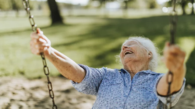 Eine ältere Frau sitzt auf einer Schaukel und lacht herzlich. Sie trägt ein blauen Hemd und hat graue Haare. Der Hintergrund zeigt eine grüne Wiese mit Bäumen im Freien, was eine fröhliche und entspannte Stimmung vermittelt.