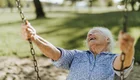Eine ältere Frau sitzt auf einer Schaukel und lacht herzlich. Sie trägt ein blauen Hemd und hat graue Haare. Der Hintergrund zeigt eine grüne Wiese mit Bäumen im Freien, was eine fröhliche und entspannte Stimmung vermittelt.