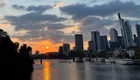 Sonnenuntergang über dem Fluss mit der Skyline von Frankfurt im Hintergrund. Die Sonne reflektiert auf dem Wasser, während Wolken den Himmel in sanften Farben erleuchten. An den Ufern sind einige Boote zu sehen.