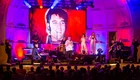 Ein Live-Konzert mit Musikern auf der Bühne, die Elvis Presley huldigen. Im Hintergrund ein großes Bild von Elvis, umrahmt von bunten Lichtern. Das Publikum ist aufmerksam und genießt die Darbietung.