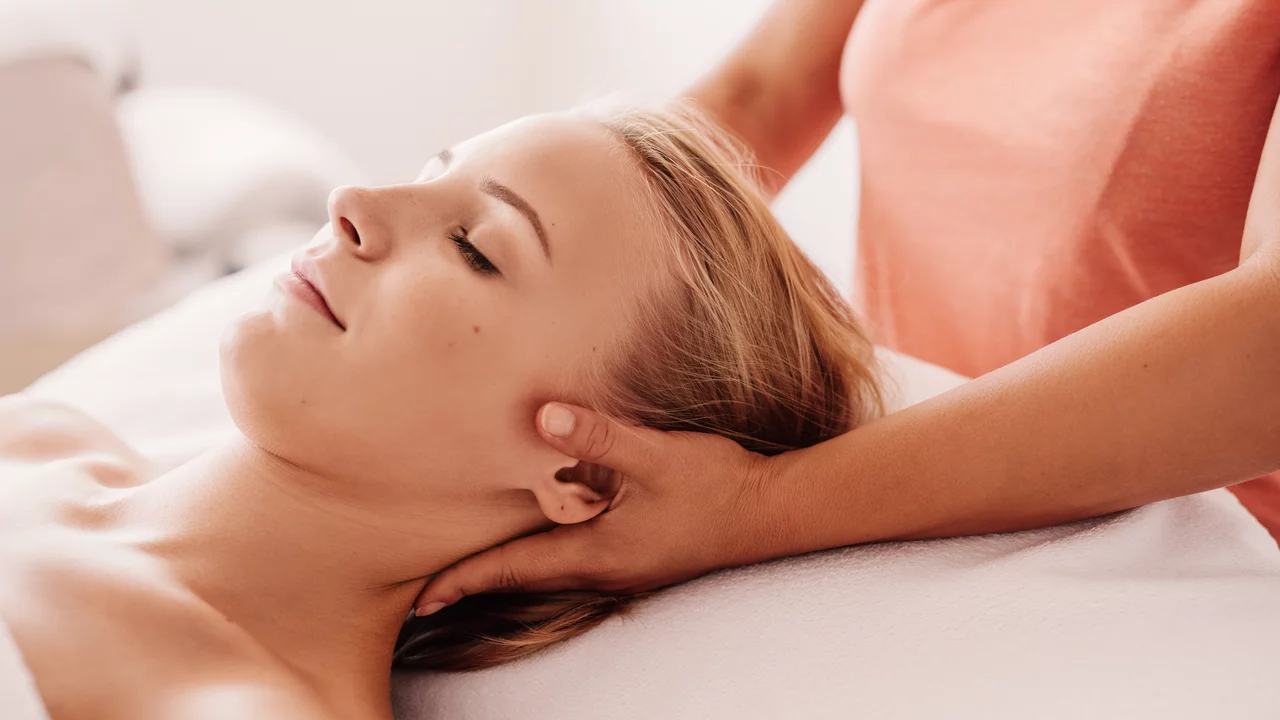 Eine Person liegt auf einer Liege und genießt eine Massage. Eine Hand übt sanften Druck auf ihren Nacken aus, während die andere Hand entspannt an ihrer Seite liegt. Die Atmosphäre wirkt friedlich und entspannend.