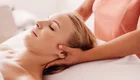 Eine Person liegt auf einer Liege und genießt eine Massage. Eine Hand übt sanften Druck auf ihren Nacken aus, während die andere Hand entspannt an ihrer Seite liegt. Die Atmosphäre wirkt friedlich und entspannend.