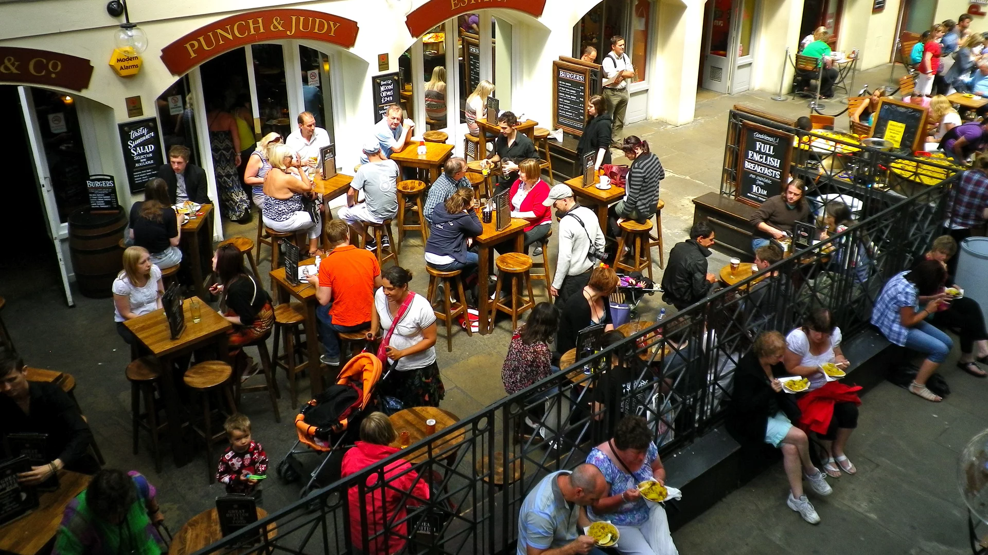 Eine belebte Gastronomie-Szene mit vielen Menschen, die an Tischen stehen oder sitzen. Einige Gäste essen und trinken, während andere miteinander sprechen. Im Hintergrund sind Außenterrassen und Eingänge zu Restaurants sichtbar. Die Atmosphäre wirkt lebhaft und gesellig.