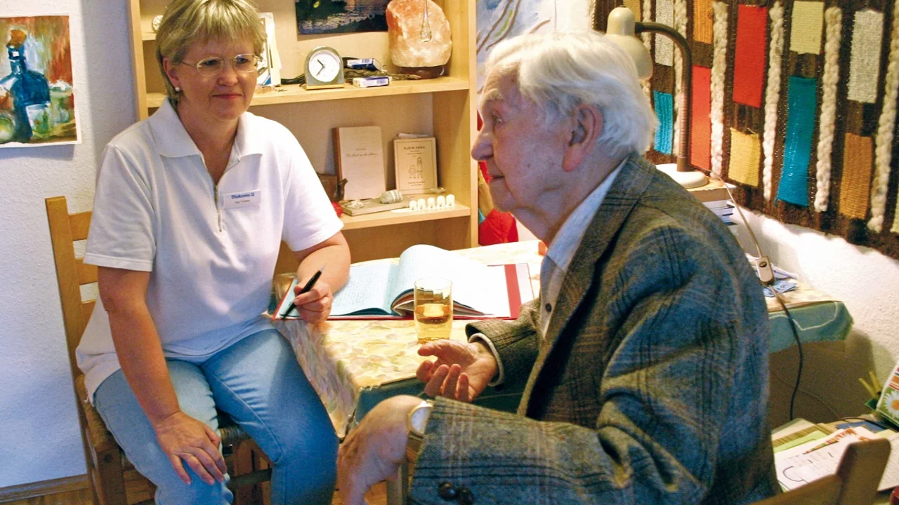 Eine ältere Dame und ein älterer Herr sitzen an einem Tisch in einem gemütlichen Raum. Die Dame, in einem weißen T-Shirt, lächelt und hält einen Stift in der Hand, während der Herr mit grauem Haar aufmerksam zuhört. Auf dem Tisch liegen Bücher und ein Glas.