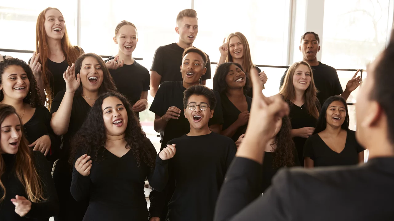 Eine Gruppe junger Menschen singt begeistert in einem Chor. Sie tragen schwarze Kleidung und zeigen verschiedene Gesichtsausdrücke, die Freude und Energie widerspiegeln. Im Vordergrund steht eine Person, die den Chor anleitet, während die anderen aufmerksam zuhören und mit Begeisterung mitsingen.