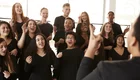 Eine Gruppe junger Menschen singt begeistert in einem Chor. Sie tragen schwarze Kleidung und zeigen verschiedene Gesichtsausdrücke, die Freude und Energie widerspiegeln. Im Vordergrund steht eine Person, die den Chor anleitet, während die anderen aufmerksam zuhören und mit Begeisterung mitsingen.