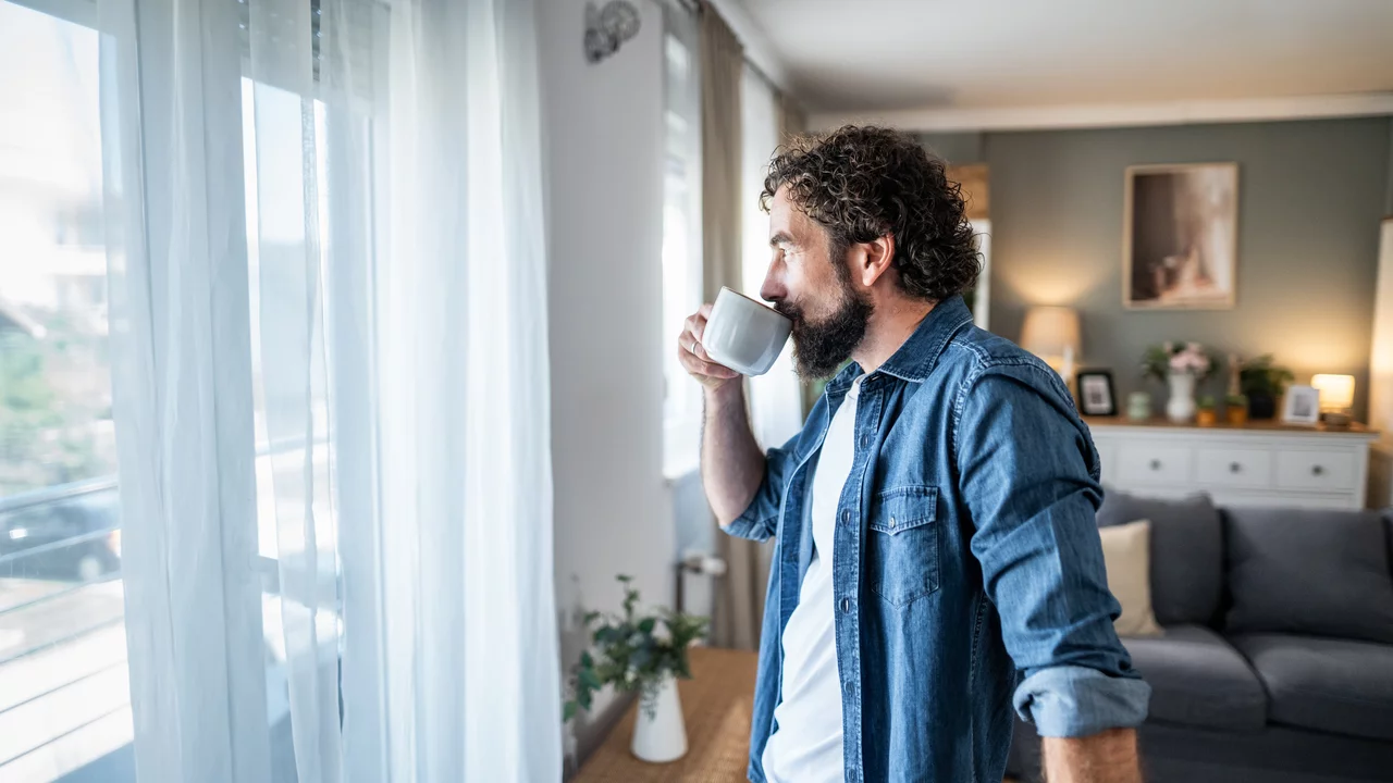 Ein Mann mit lockigem Haar steht an einem Fenster und hält eine Tasse in der Hand. Er schaut nach draußen, während sanftes Licht durch die Vorhänge strömt. Im Hintergrund sind eine Couch und dekorative Elemente zu sehen, die eine gemütliche Wohnatmosphäre schaffen.