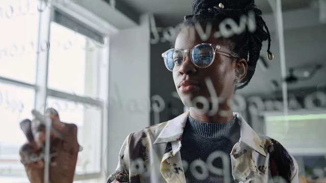 Eine Frau mit Brille schreibt mit einem Marker auf einer transparenten Oberfläche. Sie trägt ein gemustertes Hemd und schaut konzentriert auf das Geschriebene. Im Hintergrund sind unscharfe Möbel und Fenster zu erkennen.