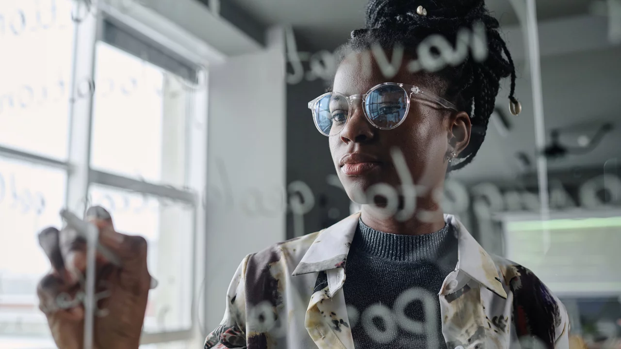 Eine Frau mit Brille schreibt mit einem Marker auf einer transparenten Oberfläche. Sie trägt ein gemustertes Hemd und schaut konzentriert auf das Geschriebene. Im Hintergrund sind unscharfe Möbel und Fenster zu erkennen.