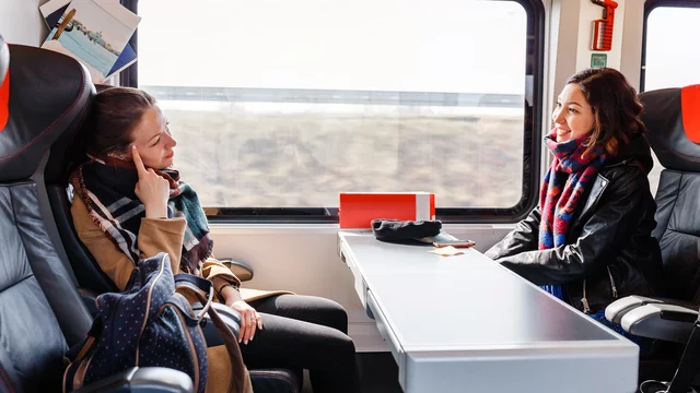 Zwei Frauen sitzen im Zug gegenüber an einem Tisch. Die eine lehnt sich zurück und schaut nachdenklich, während die andere entspannt in ihre Richtung lächelt. Beide tragen warme Kleidung und eine gemütliche Atmosphäre ist spürbar. Durch das Fenster sieht man die vorbeiziehende Landschaft.