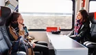 Zwei Frauen sitzen im Zug gegenüber an einem Tisch. Die eine lehnt sich zurück und schaut nachdenklich, während die andere entspannt in ihre Richtung lächelt. Beide tragen warme Kleidung und eine gemütliche Atmosphäre ist spürbar. Durch das Fenster sieht man die vorbeiziehende Landschaft.